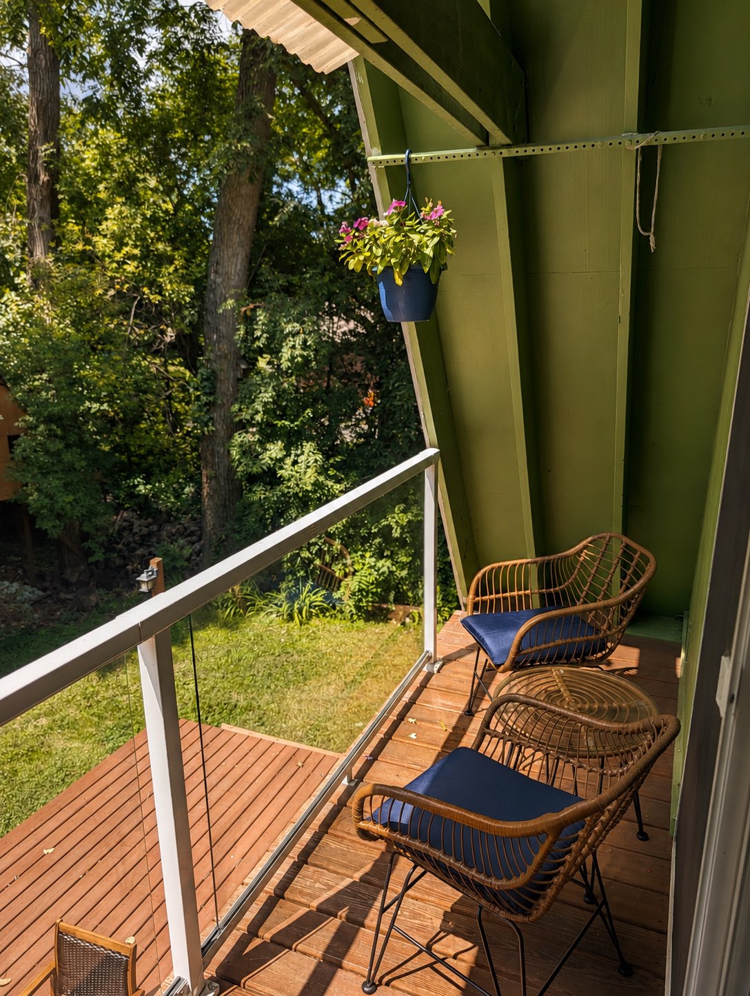 The upper balcony deck under the green A-frame eaves, with rattan chairs, a hanging flower basket, and a cable railing looking into the trees.