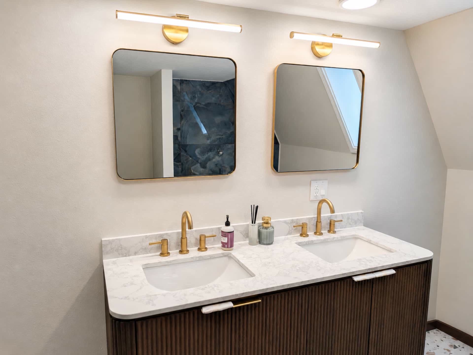 Bathroom with a double vanity, two mirrors, gold fixtures, and a fluted dark cabinet under a sloped ceiling.