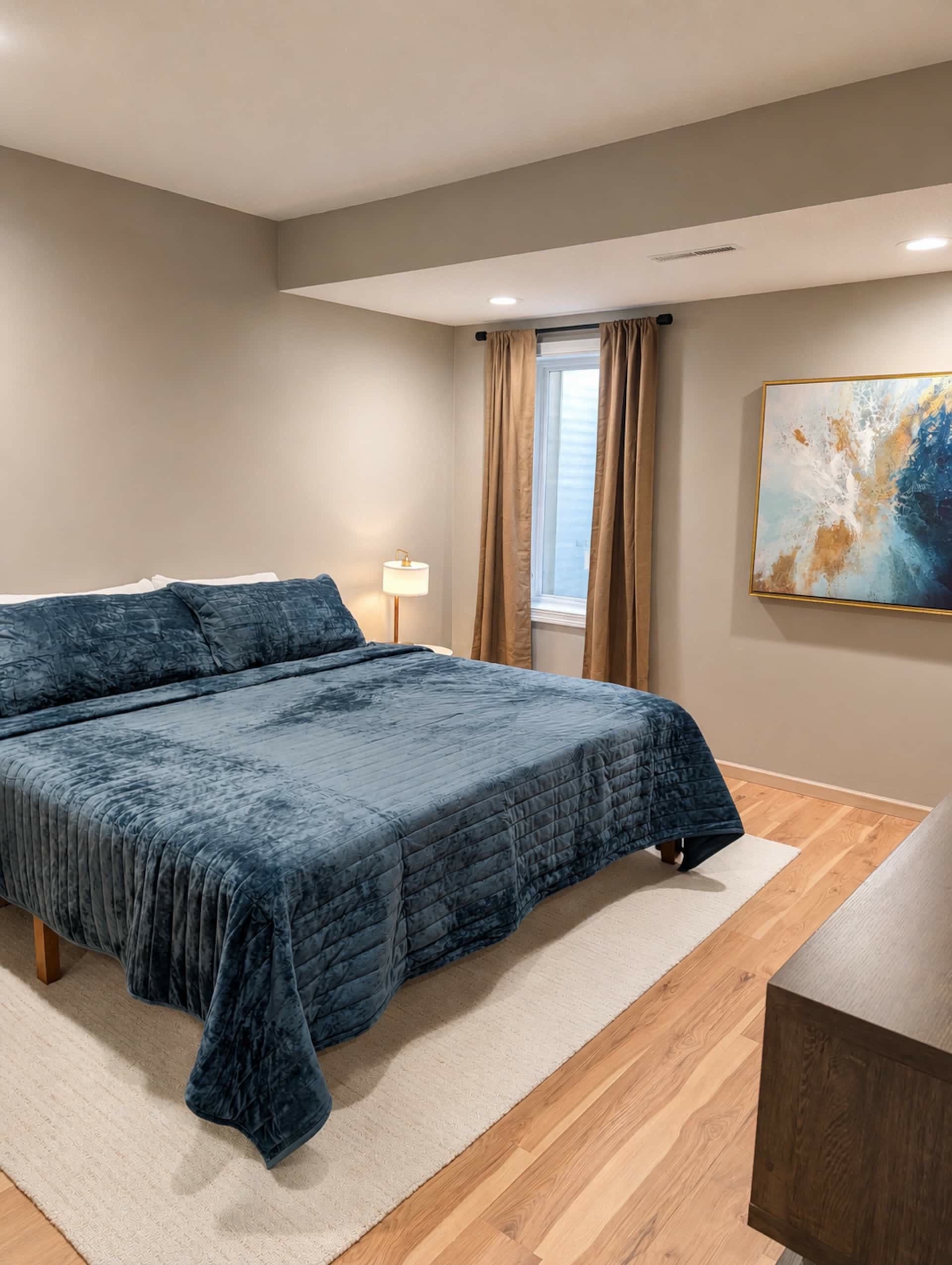 Bedroom with a king bed dressed in a blue velvet throw on a wood platform, framed art, and a bright window.