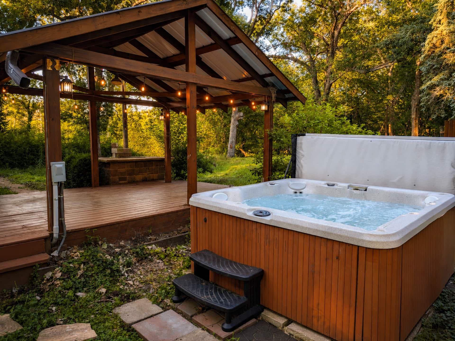 The private hot tub beside a covered patio strung with warm lights, surrounded by green trees at golden hour.