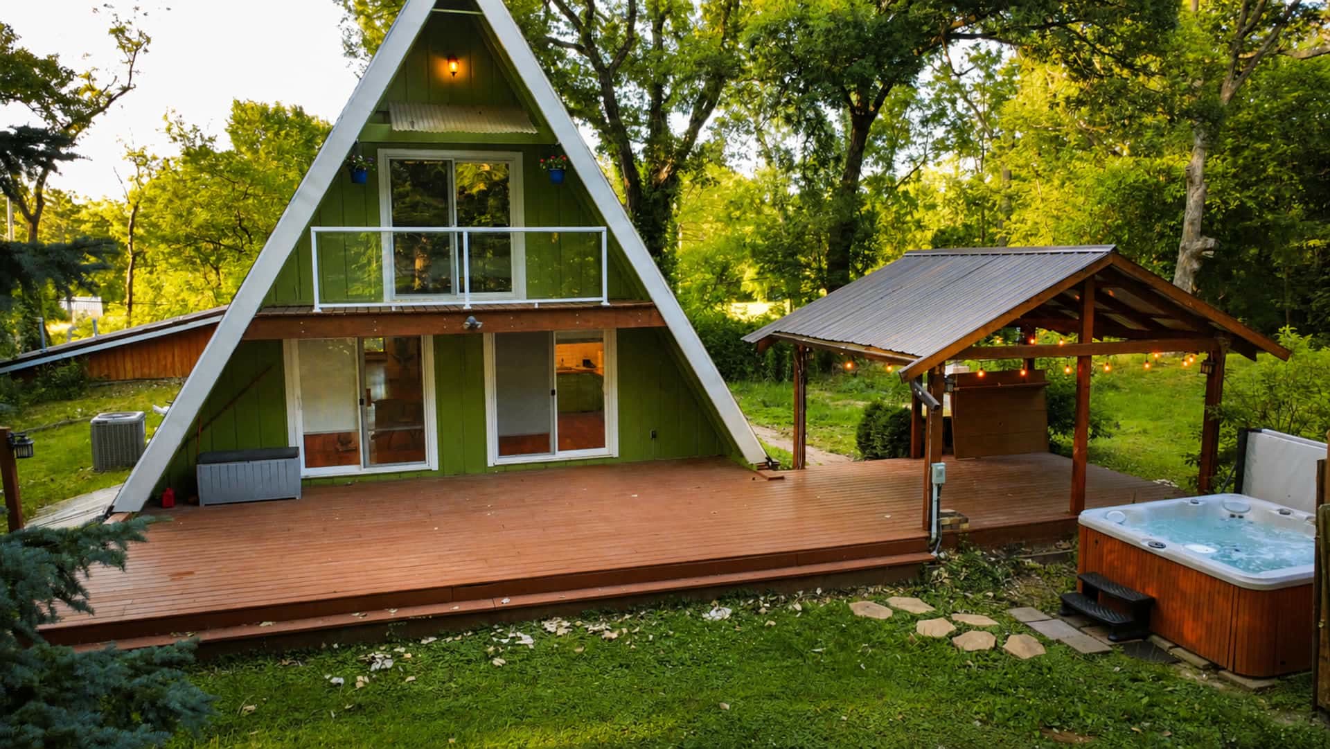 The green A-frame at dusk with a long wood deck, a covered gazebo strung with lights, and a private hot tub, surrounded by trees.