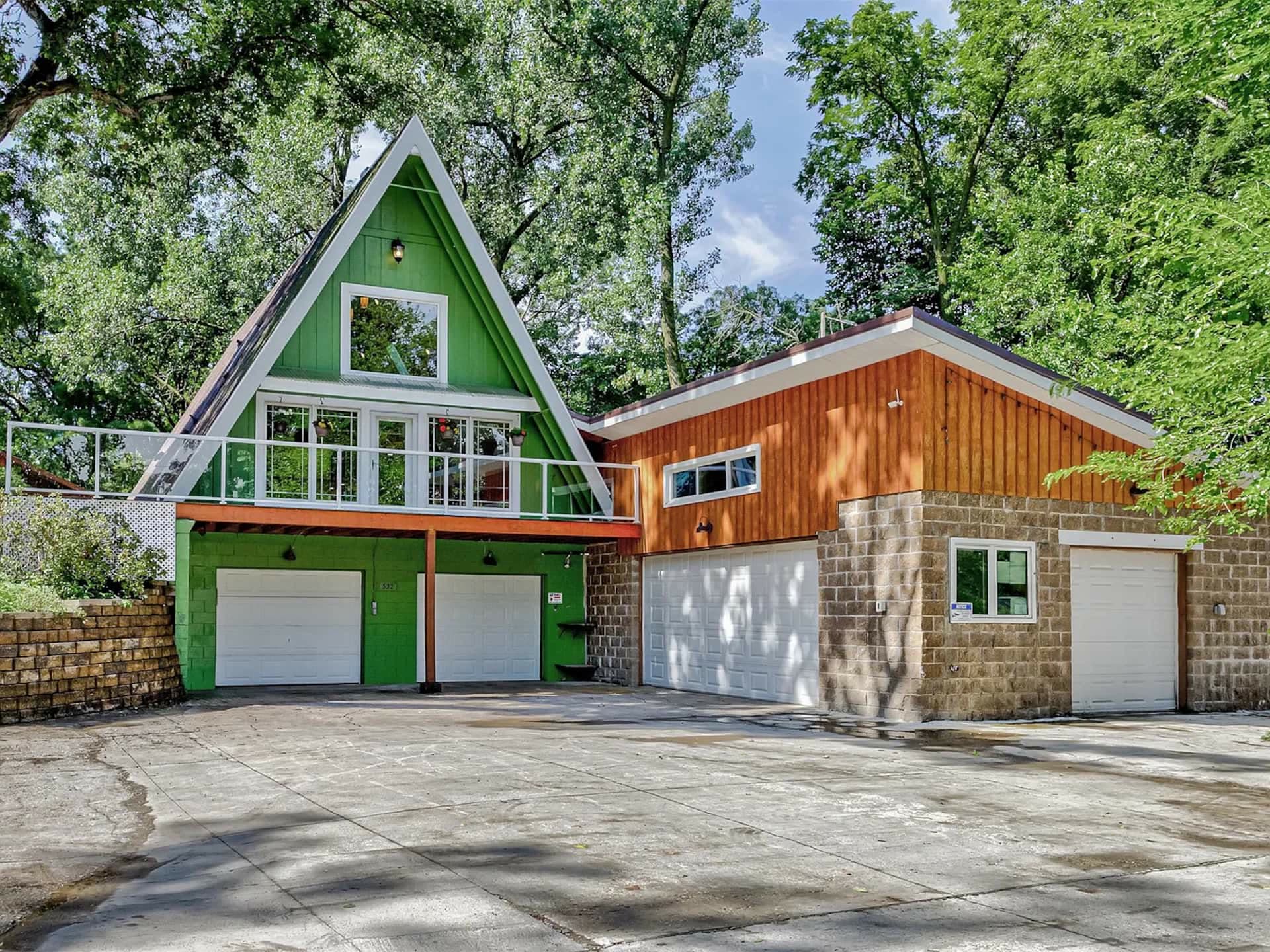 The green A-frame house with white-trimmed peak, a balcony and wall of windows, set among tall trees.
