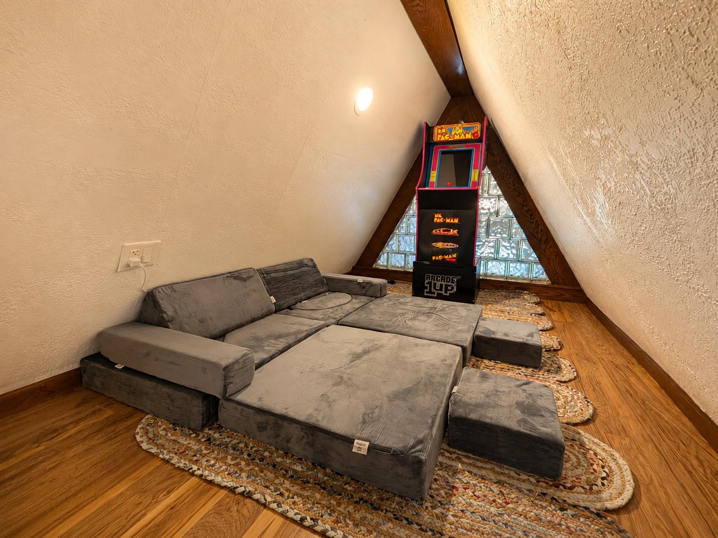 A children's loft under the A-frame peak with a gray modular play couch, braided rugs, and a retro Ms. Pac-Man arcade cabinet by a glass-block window.