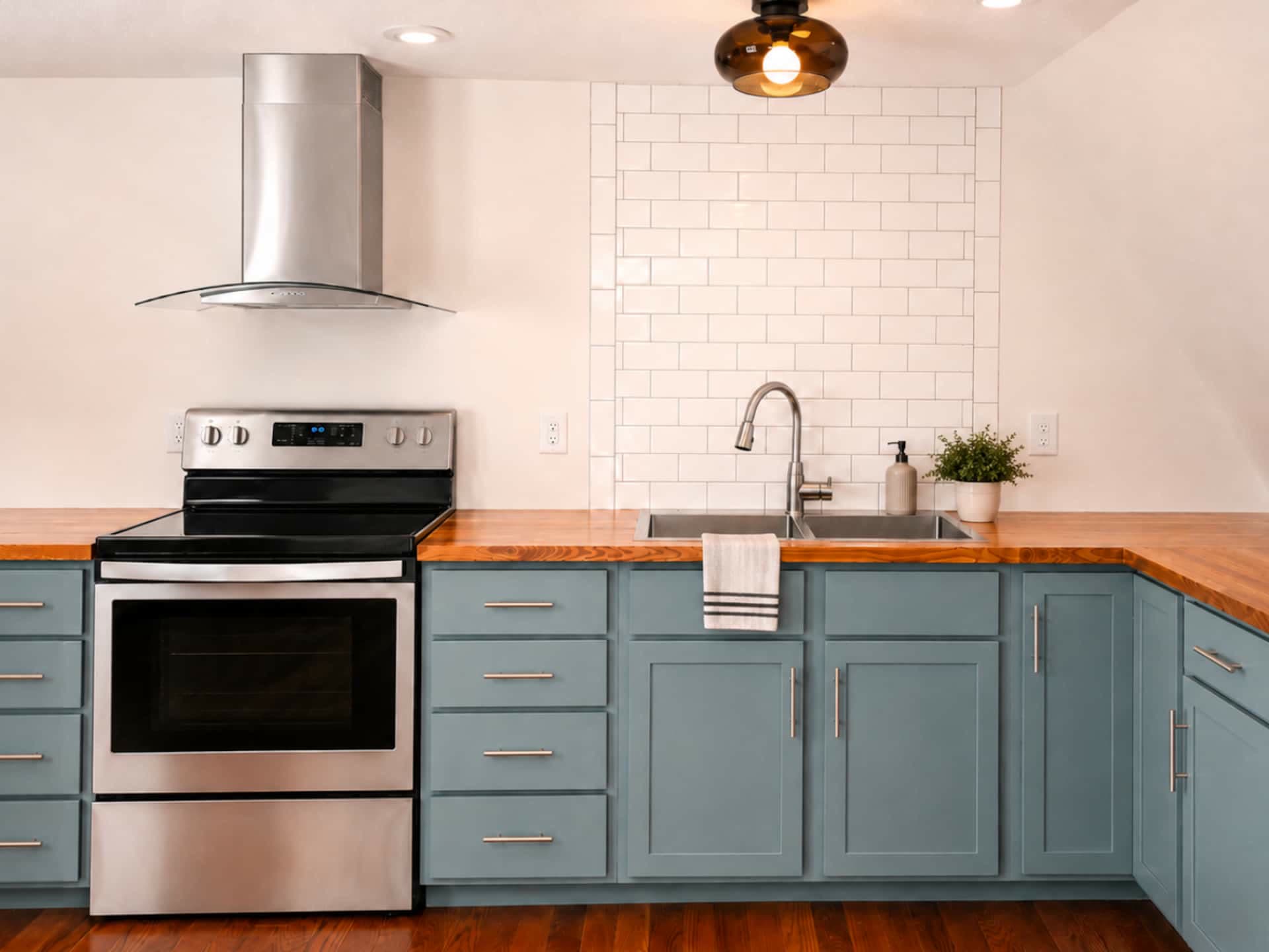Full kitchen with a stainless range and hood, butcher-block wood counters, sage-green cabinets, and white subway tile.