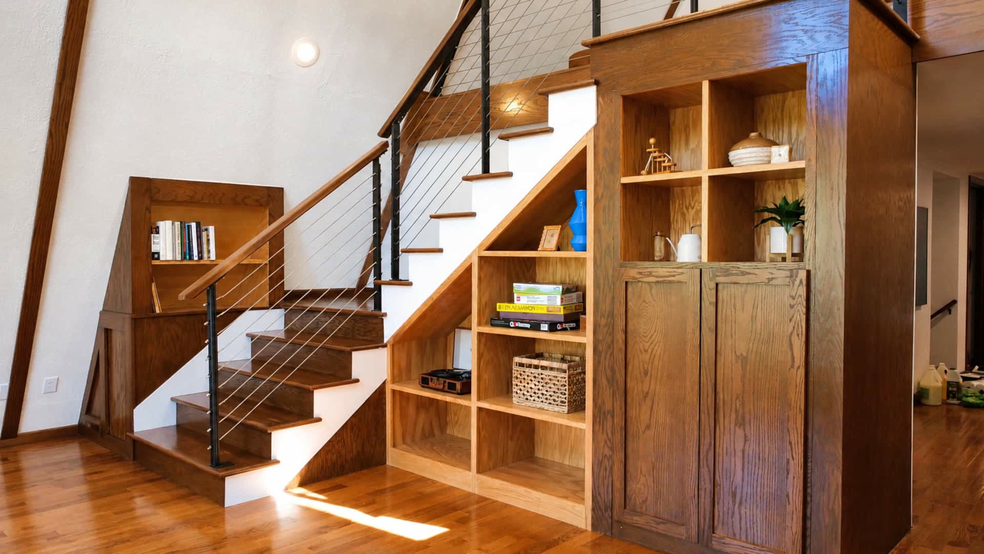 A cable-railed staircase beside a built-in red oak bookshelf and cabinet, with wood floors under the sloped ceiling.