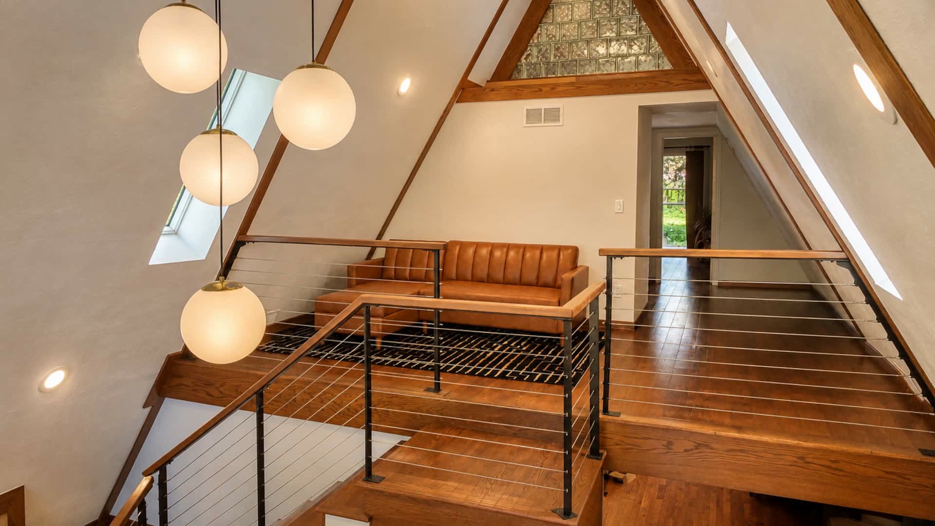 Upper landing with globe pendant lights, a tan leather bench, cable railing, and exposed A-frame beams with skylights.