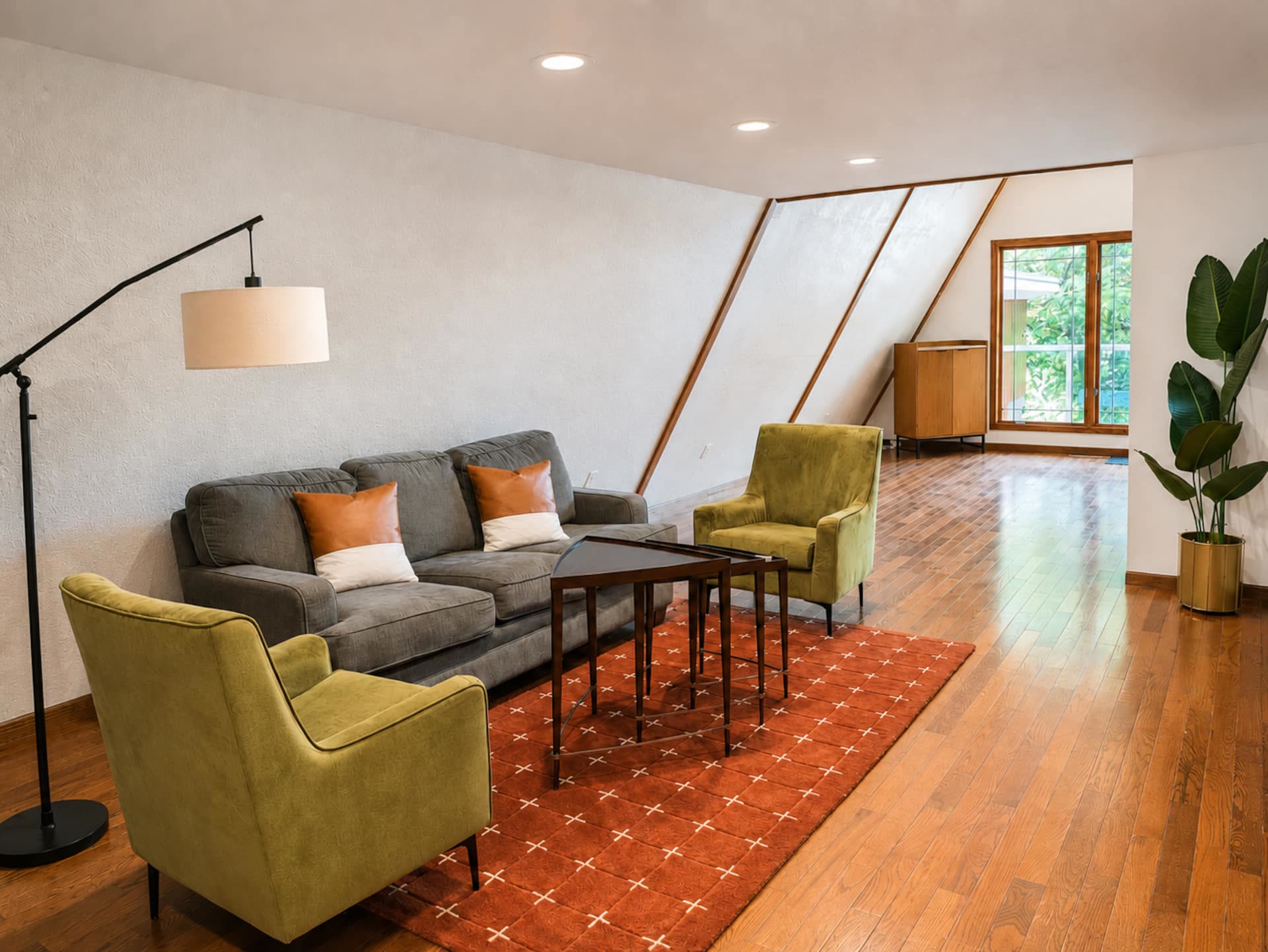 A-frame living room with a sloped ceiling, exposed wood beams, warm red oak finishes, a gray sofa, olive velvet armchairs, and a large window facing the trees.