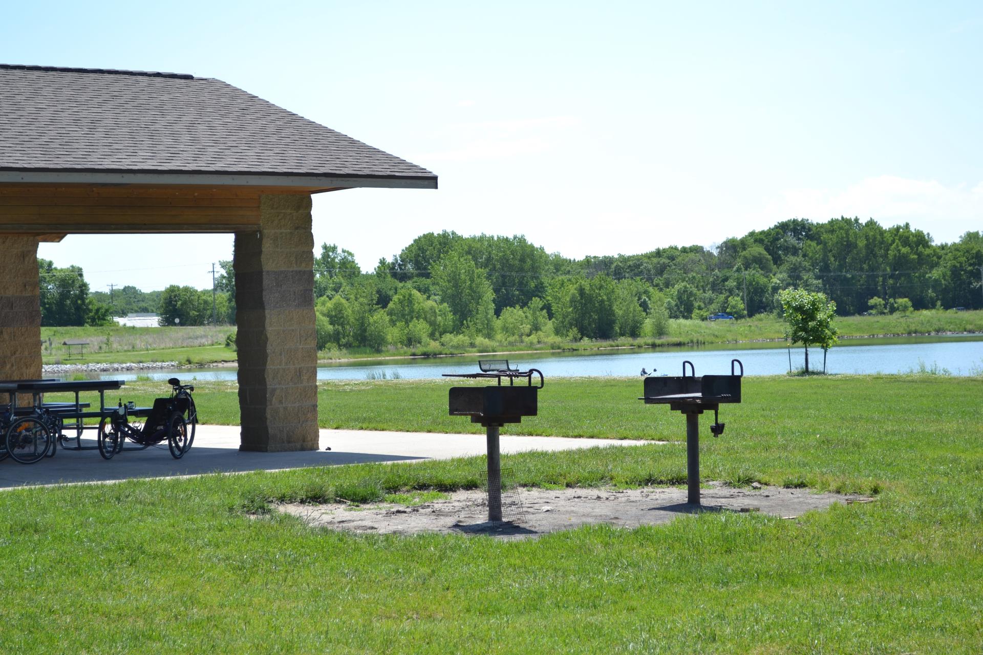 Charcoal grills on a lawn beside the paved trail, with the open lake and wooded far shore behind.