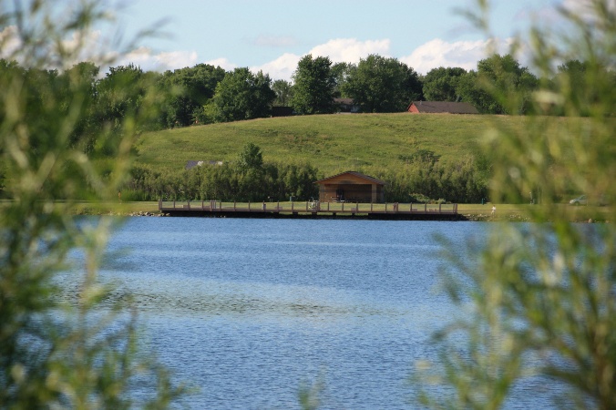 Ada Hayden Lake framed by willow branches, with a fishing dock and picnic shelter across the water under a blue sky.