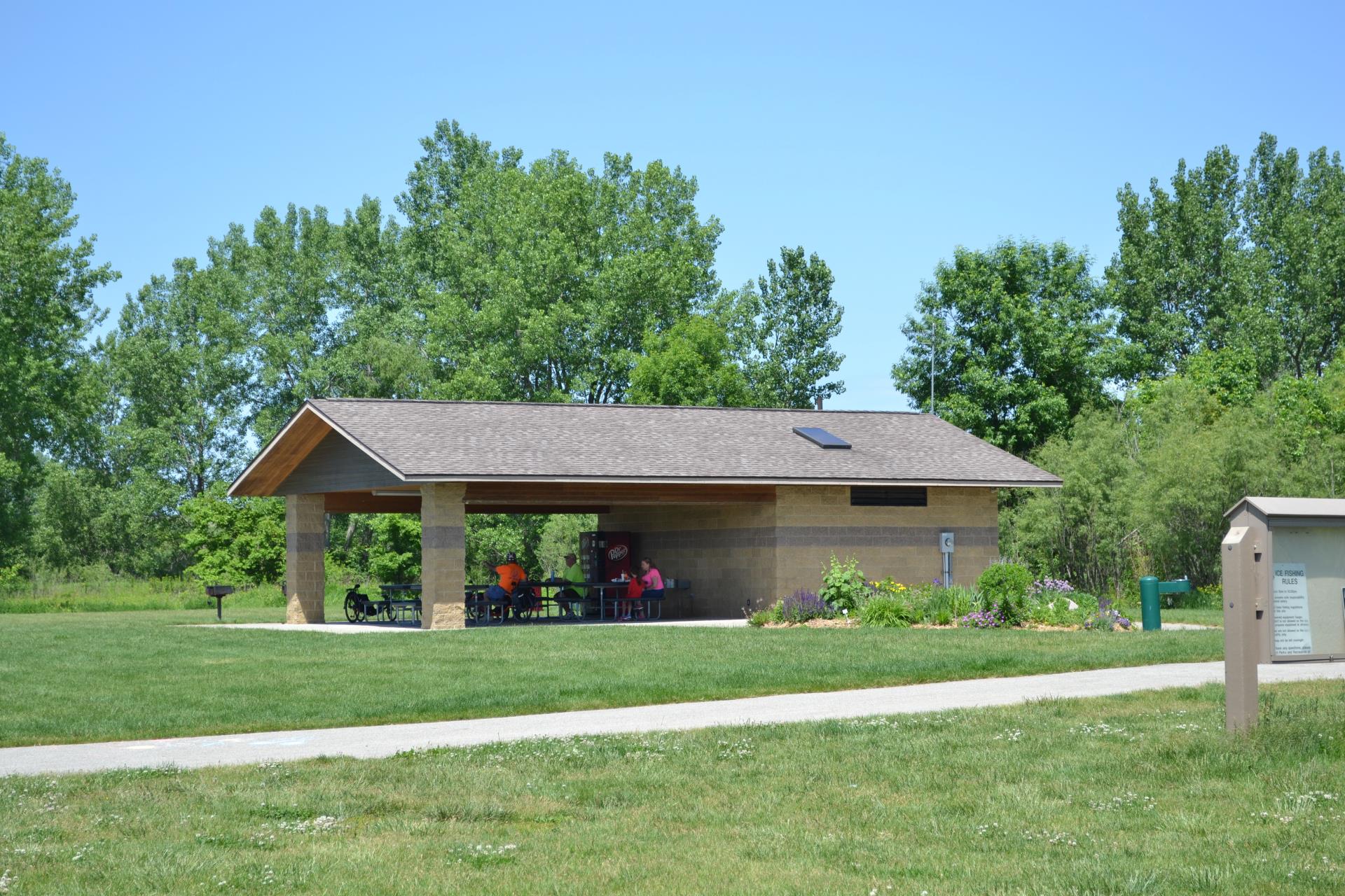 The park's covered picnic shelter with tables, a paved path, and prairie plantings on a sunny day.